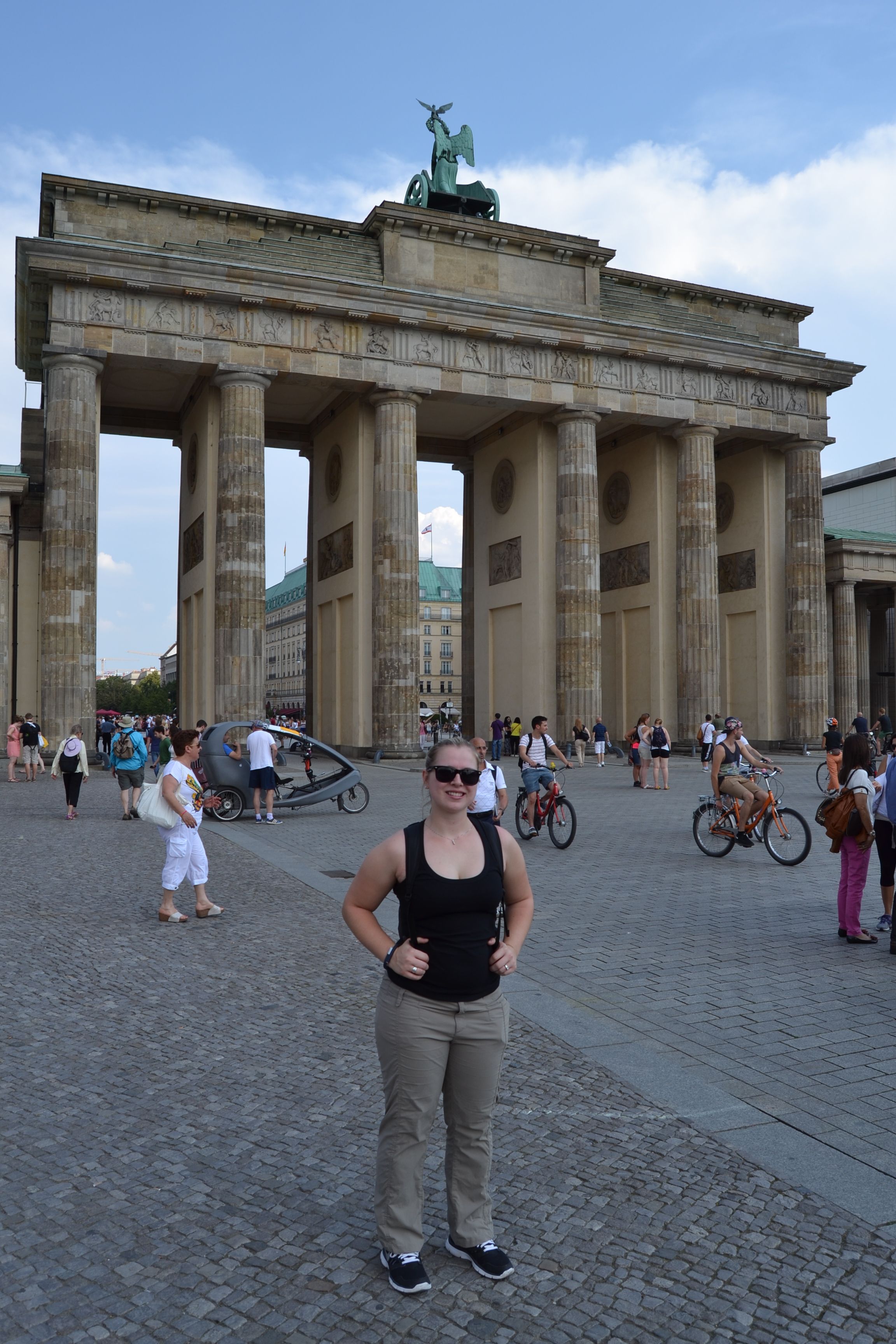 white woman in tan pants, black tank top and wearing sunglasses standing in front of the Brandenburg Gate