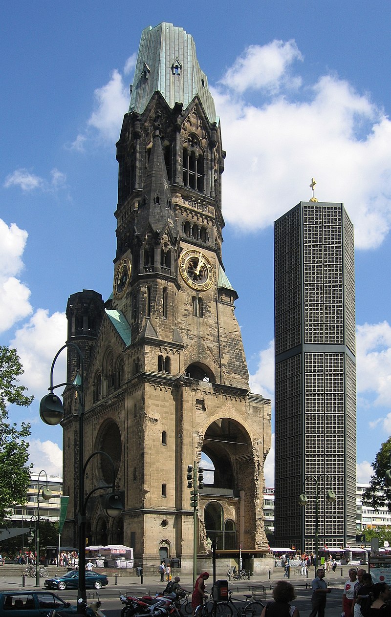 Image of the Kaiser Wilhelm Memorial Church - a large brick building that shows signs of damage from conflicts against a bright blue sky with clouds